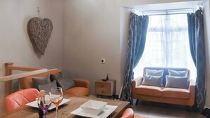 A living room with a wooden dining table, orange chairs, a sofa with cushions, a heart-shaped wall decoration, and a window with curtains. - 1 bedroom accommodation in Windermere (Windermere)