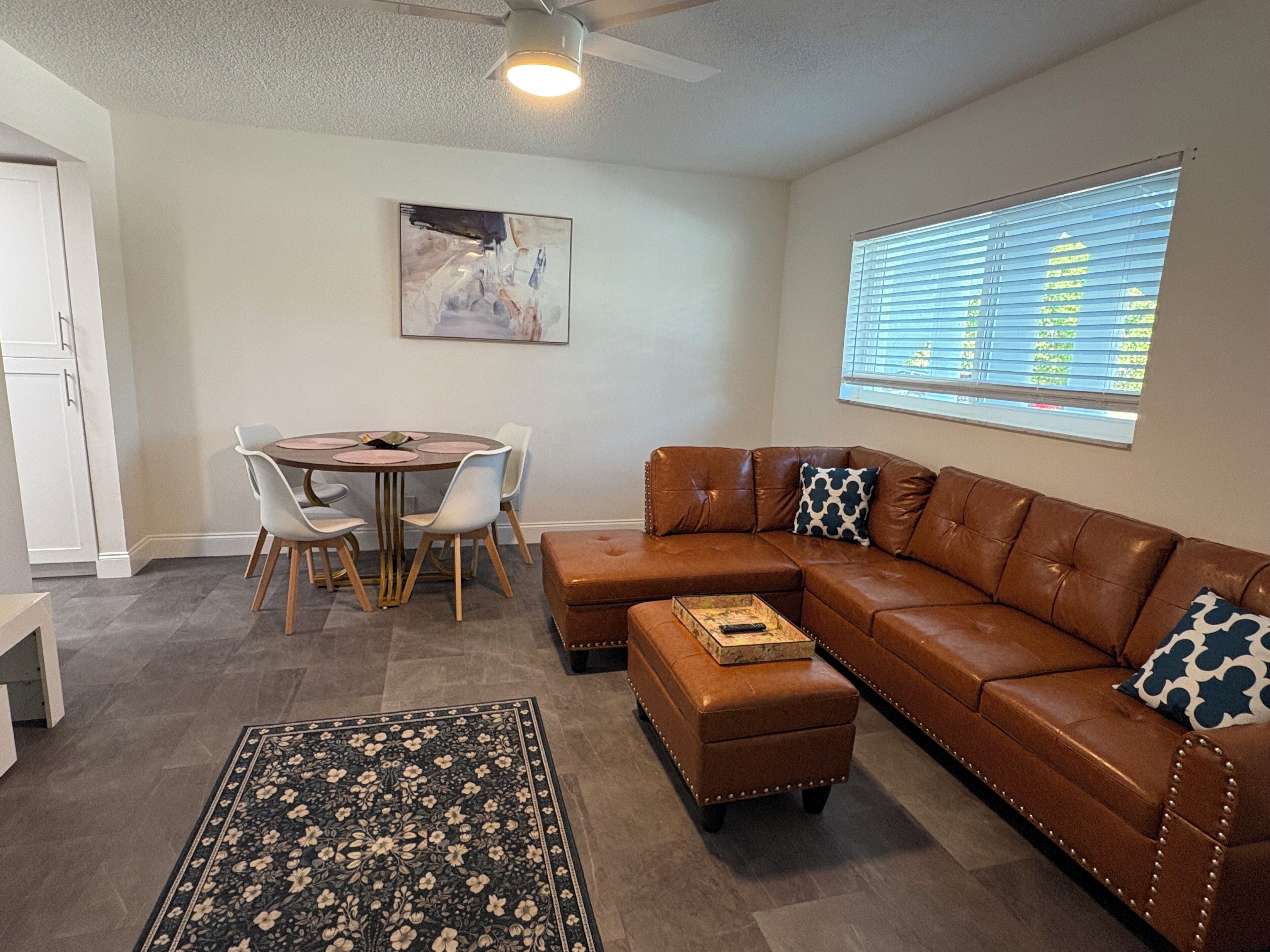 A living room with a brown leather sectional sofa, a round dining table with chairs, a coffee table, a patterned rug, and a ceiling fan.