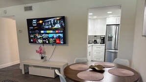 Ein modernes Wohnzimmer mit an der Wand montiertem Fernseher, der verschiedene Streaming-Dienste anzeigt, ein Essbereich mit rundem Tisch und Stühlen sowie eine Küche mit weißen Schränken und Edelstahlgeräten.