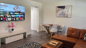 Ein modernes Wohnzimmer mit einem Flachbildfernseher, der eine Streaming-Dienstanwendung anzeigt, einem Essbereich mit rundem Tisch und Stühlen, einem braunen Ledersofa und einem dekorativen Teppich.