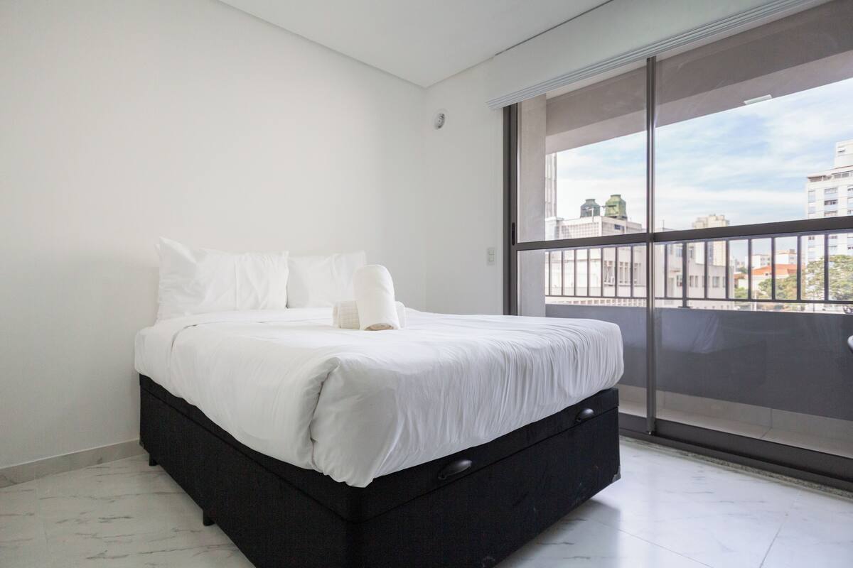 Un dormitorio moderno con una cama grande, un cabecero de color oscuro y vista a la ciudad a través de un ventanal amplio.