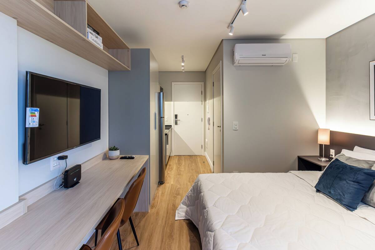 Una habitación de hotel moderna con una televisión de pantalla plana grande, un escritorio con computadora, una cama con ropa de cama blanca y una mesita de noche con lámpara.