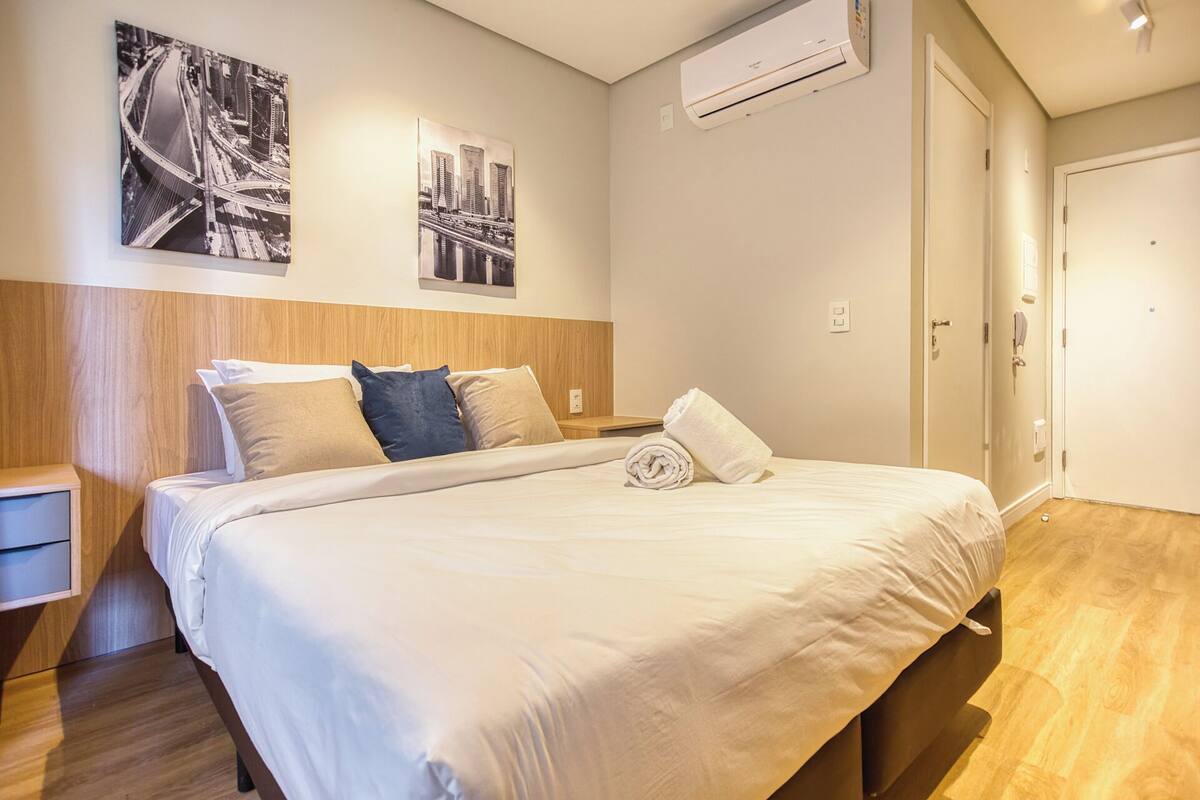 Una habitación de hotel con cama, mesita de noche, unidad de aire acondicionado y dos fotografías en blanco y negro de un paisaje urbano en la pared.