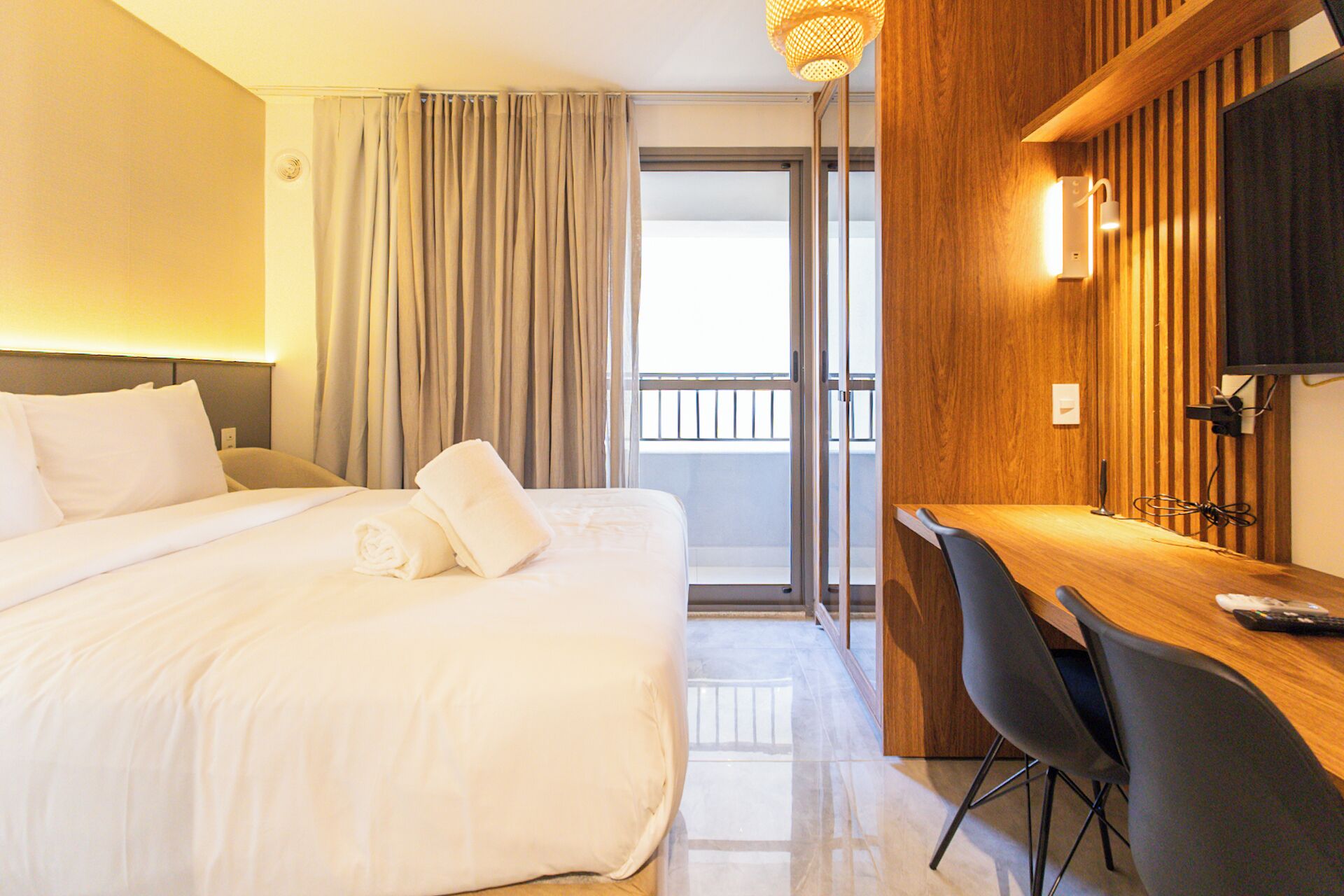 A hotel room with a bed, a desk with a chair, a television, and a balcony with curtains.