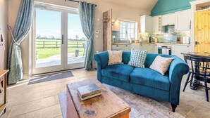 A living room with a blue sofa, a wooden coffee table with books, a kitchen in the background, and a view of a green field through the glass door. - 3 bedroom accommodation in Wedmore, near Glastonbury (Wedmore, near Glastonbury)