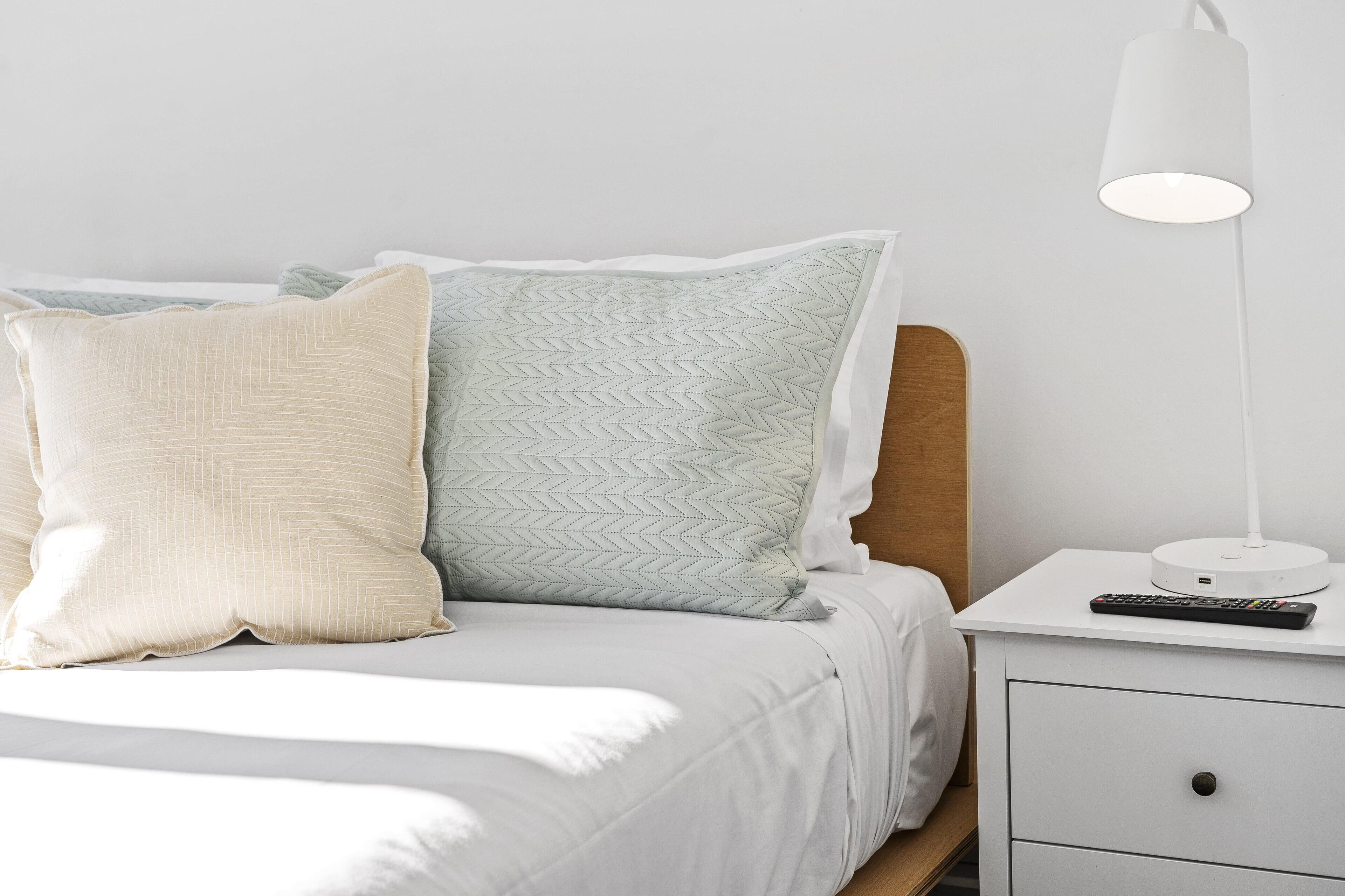 Una cama bien tendida con dos almohadas, una mesita de noche con una lámpara y un control remoto.