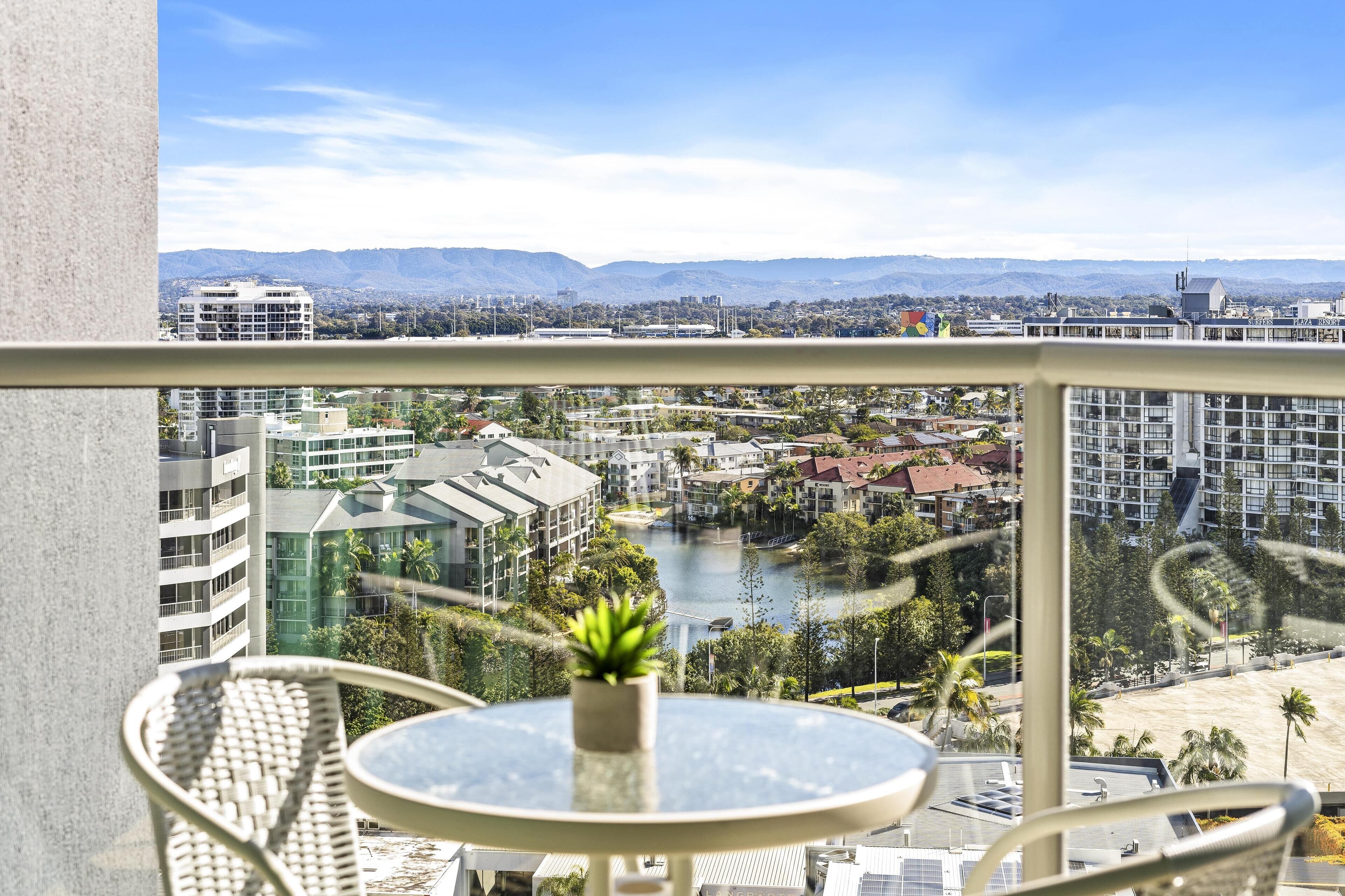 Ein Balkon mit Glastisch und Stühlen, mit Blick auf eine Stadtlandschaft mit Gebäuden, einem Fluss und Bergen.