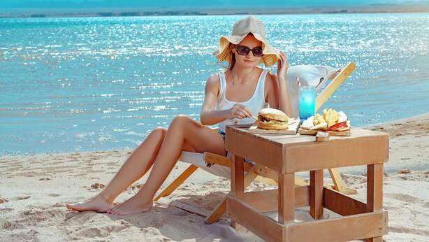 A person sitting on a beach chair with a table beside them, holding a burger and a drink. - The Kaktus Somabay (Soma Bay)