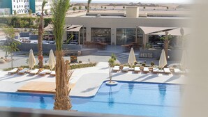 Un espace piscine avec des chaises longues et des parasols, un restaurant doté d’une terrasse, et une vue sur le désert en arrière-plan.