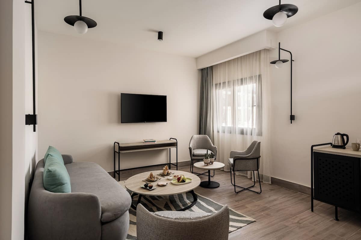 A modern living room with a sofa, two armchairs, a coffee table, a TV mounted on the wall, and a dining area with a small table and chairs.
