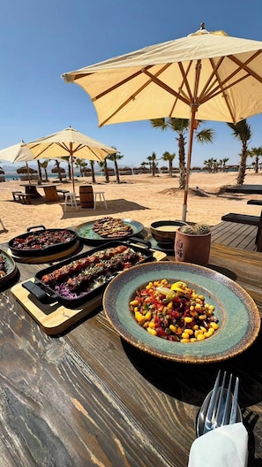 Eine Holzterrasse mit Essplätzen im Freien, Sonnenschirmen und verschiedenen Speisen, darunter gegrilltes Fleisch und ein Salat.