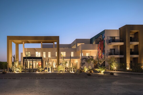 Un edificio moderno con ingresso in vetro, circondato da piante desertiche e un grande murale.