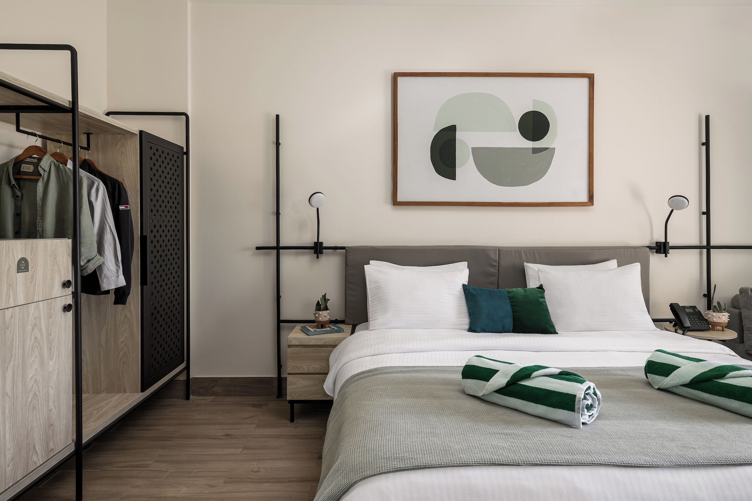 A modern hotel room with a neatly made bed, a wardrobe, a bedside table with a lamp, and a framed abstract artwork on the wall.
