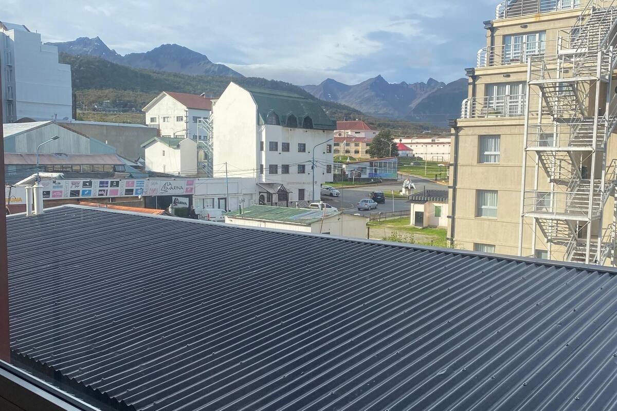 A view from a window with a corrugated metal roof, overlooking a town with buildings, a green hill, and mountains in the distance.