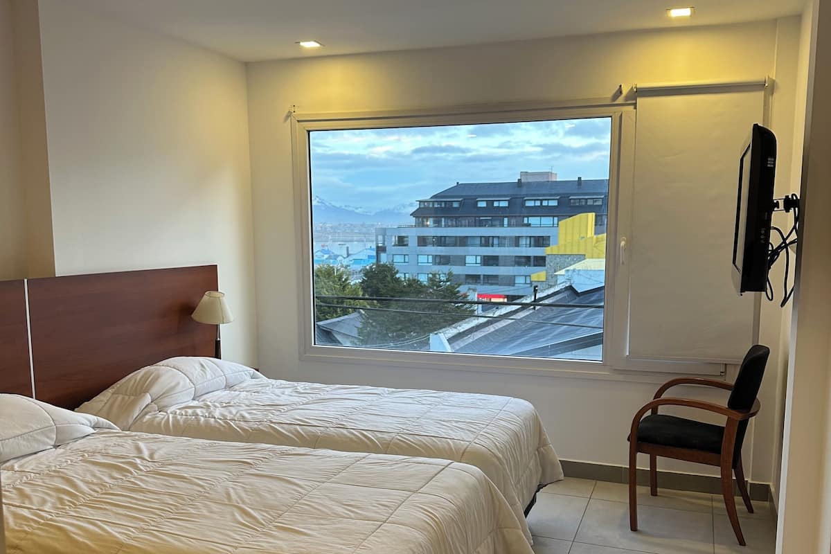 A hotel room with two beds, a window with a view of buildings and mountains, a chair, and a wall-mounted TV.