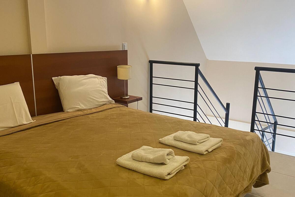 A hotel room with a large bed, a nightstand, a lamp, and a view of the outside through a window.