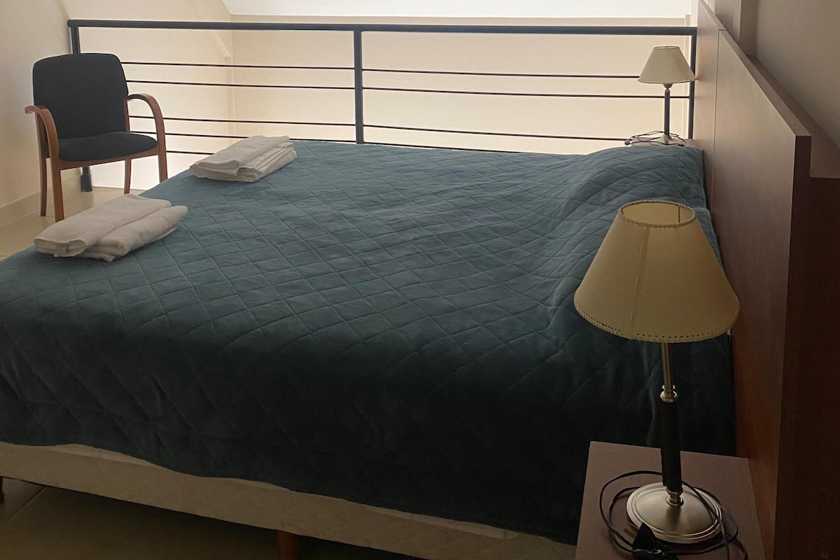 A bedroom with a bed, a chair, a lamp, and a view of the sky through a window.