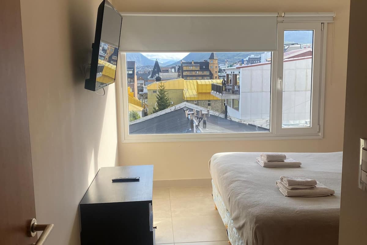 A bedroom with a bed, a desk, a TV mounted on the wall, and a window with a city view.