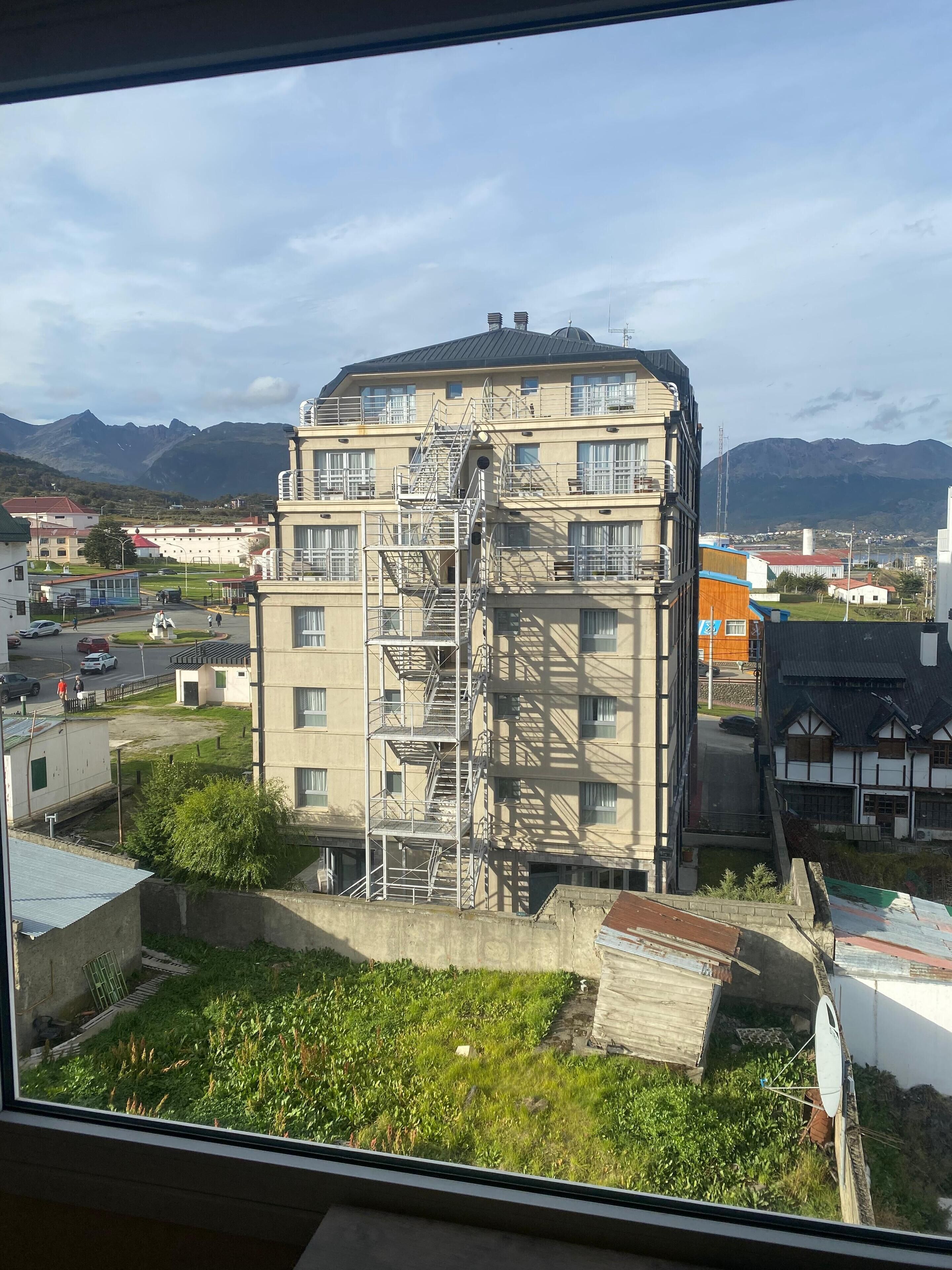 Un edificio de varios pisos con una escalera exterior destacada, rodeado de vegetación y otros edificios.