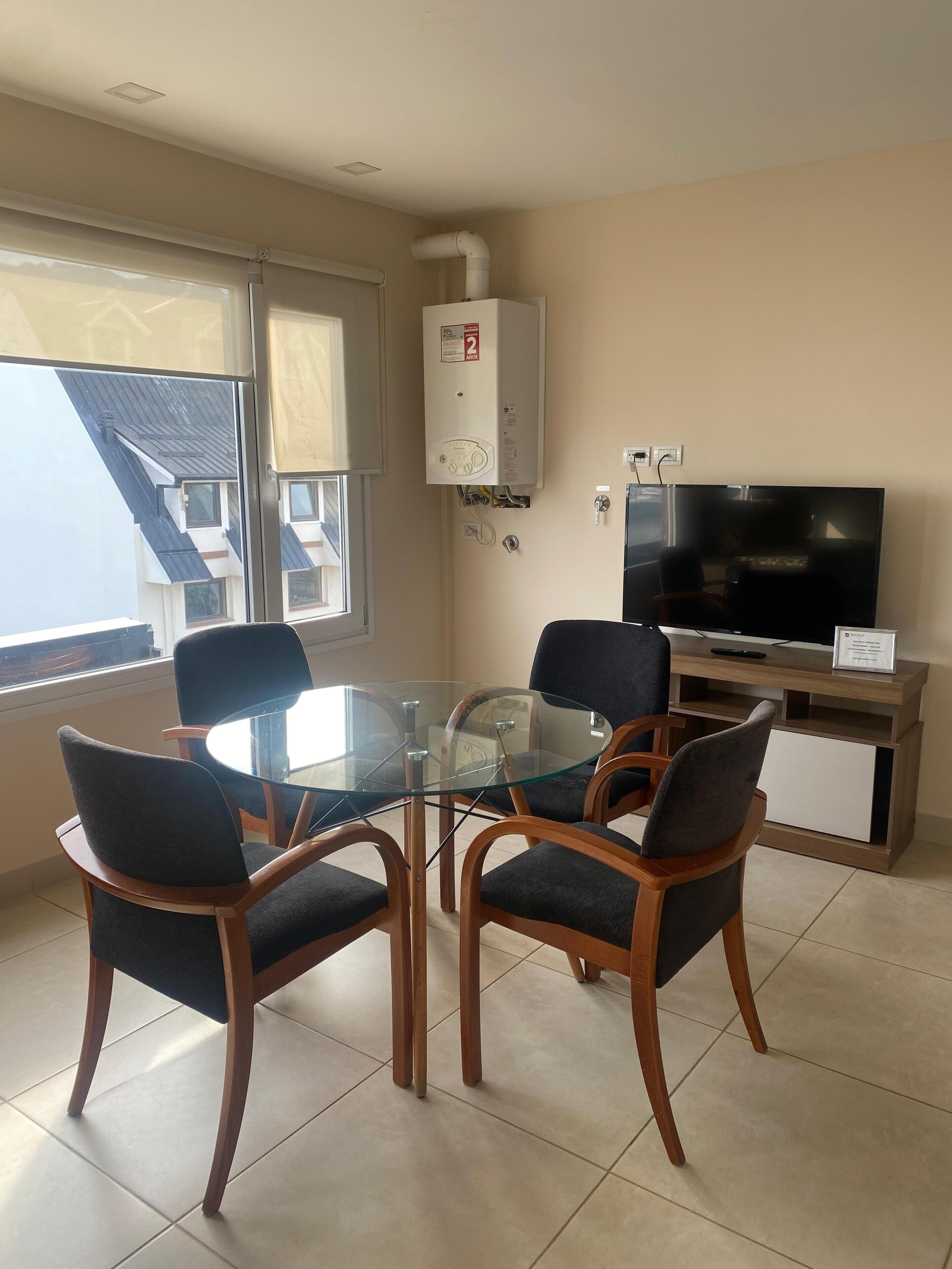 A modern living room with a glass-top dining table surrounded by four chairs, a flat-screen TV on a wooden stand, and a white radiator on the wall.