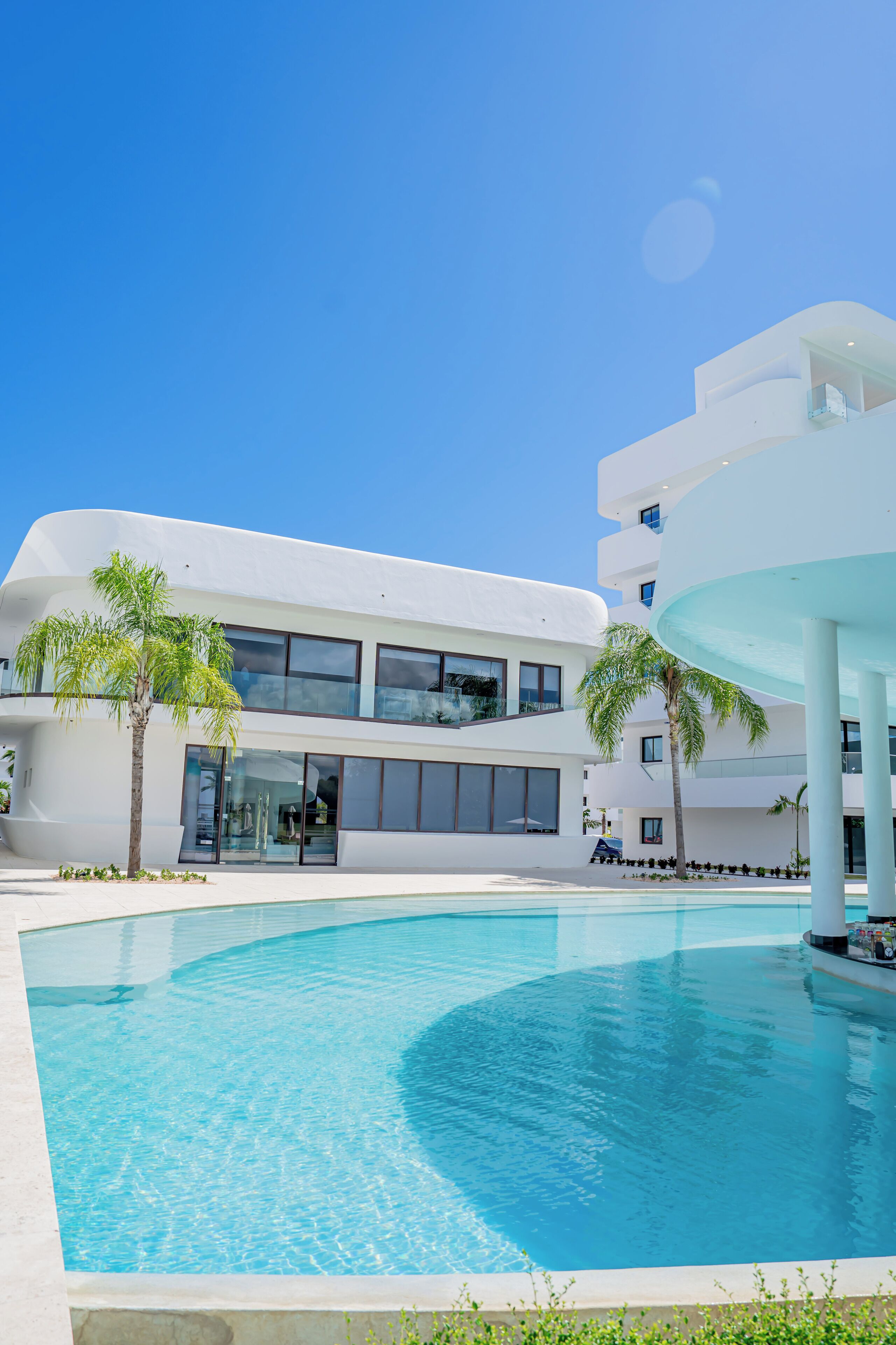 Un bâtiment moderne doté d’une piscine, de palmiers et surplombant un ciel d’un bleu éclatant.