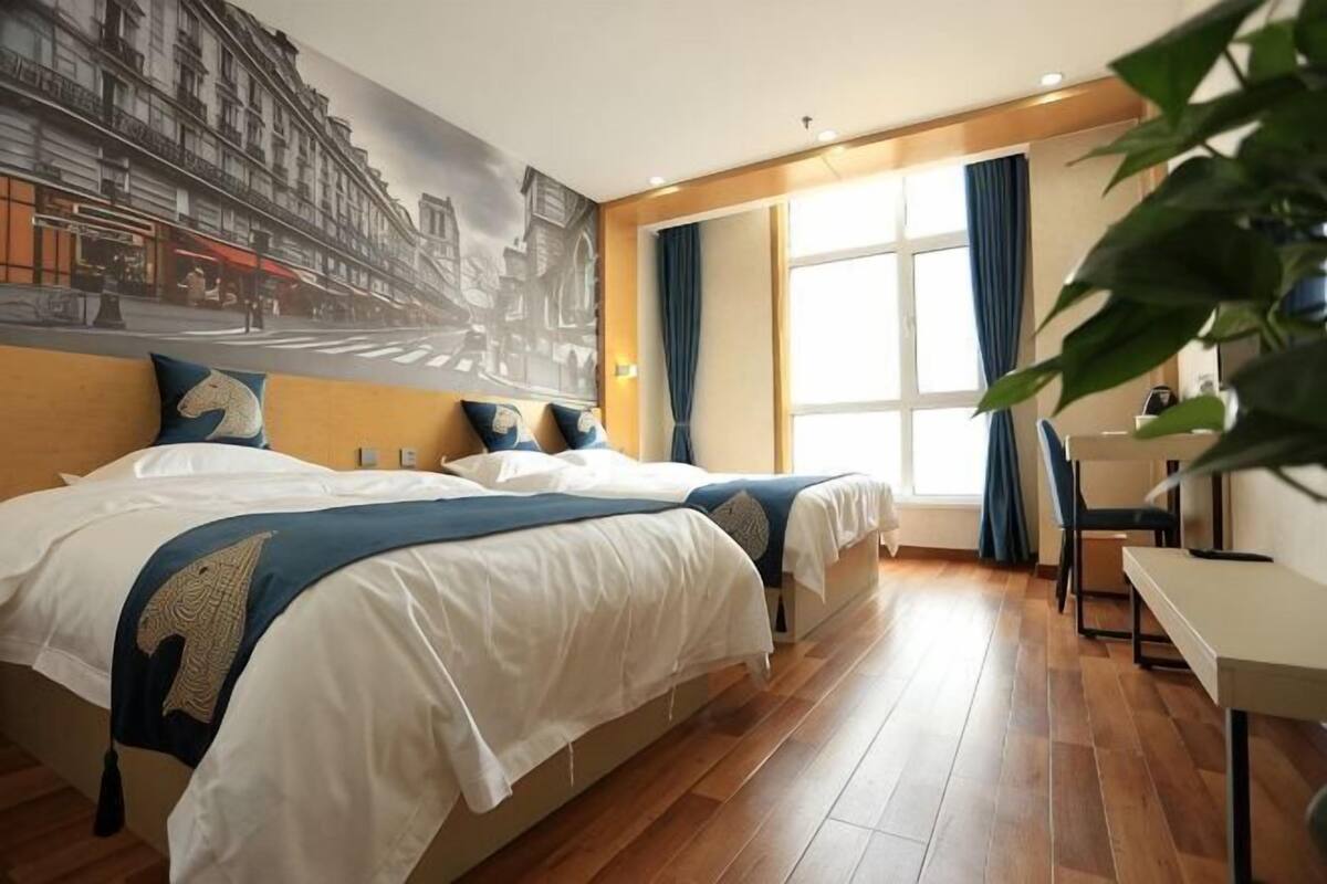 A hotel room with two beds, a desk, a chair, a large window with curtains, and a mural on the wall.