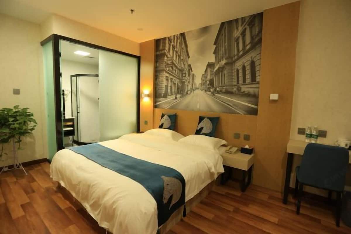 A hotel room with a bed, a desk, a chair, a plant, and a large cityscape painting on the wall.