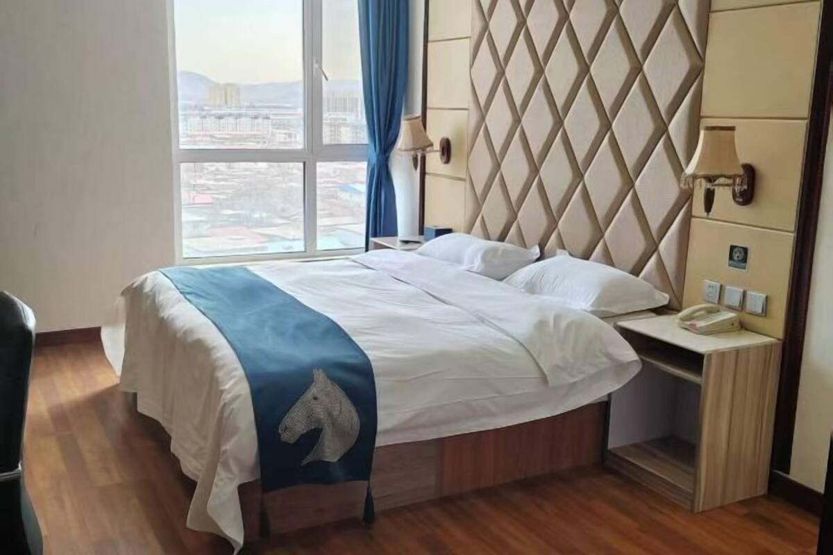 A hotel room with a bed, a nightstand, a lamp, and a view of the city.