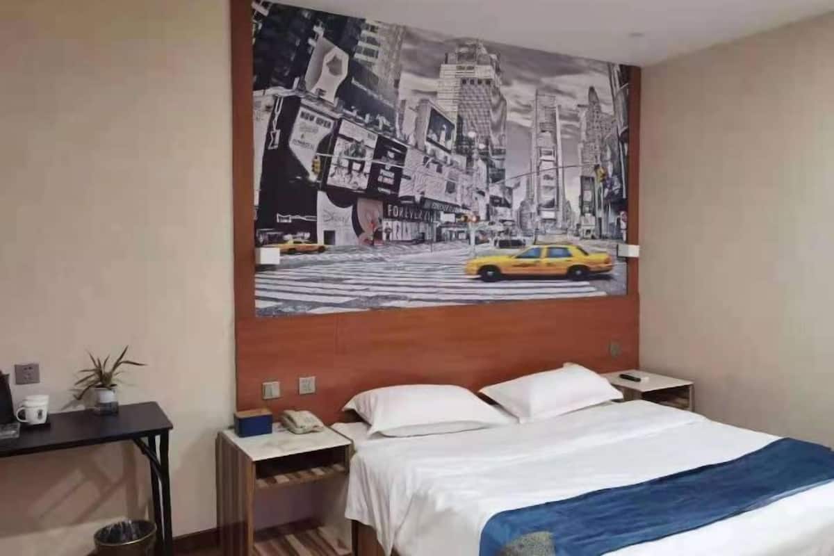 A hotel room with a bed, a nightstand, a desk, a wall-mounted telephone, and a wall tapestry depicting a cityscape.