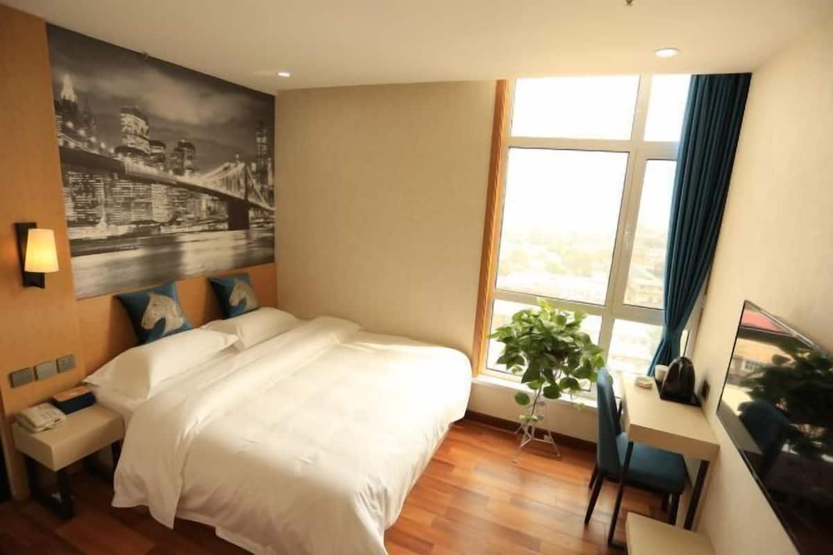 A hotel room with a bed, a desk, a chair, a window with curtains, a plant, and a cityscape painting on the wall.