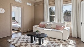 Ein Wohnzimmer mit einer Couch, einem Couchtisch und einem Teppich. Ein Fenster mit Vorhängen, eine Uhr an der Wand und eine Tür zu einem anderen Raum sind ebenfalls zu sehen.