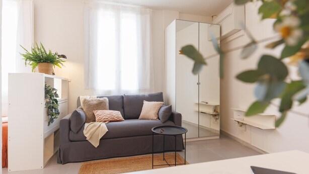 Un salon moderne avec un canapé gris, une petite table ronde et une grande fenêtre ornée de rideaux légers.