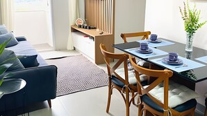 Una sala moderna con un sofá, una mesa de comedor dispuesta con tazas y platillos azules, una pequeña mesita de madera con una lámpara y una planta en maceta.