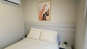 Una cama bien hecha con sábanas blancas, un cabecero, una mesita de noche con una planta pequeña y un aire acondicionado fijado a la pared.