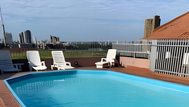 A rectangular swimming pool with a metal ladder in the foreground, surrounded by white lounge chairs and a brick deck. - APTO decorado 5 hospedes AC e piscina (Foz do Iguaçu)