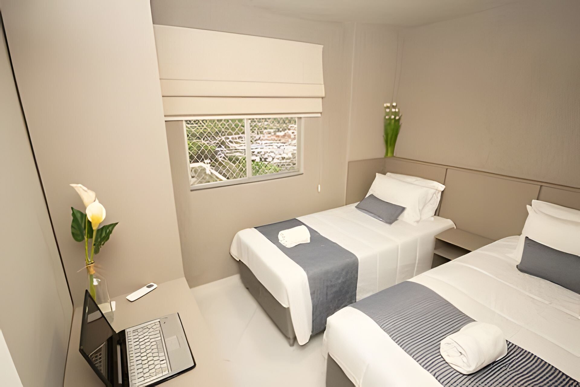 Habitación de hotel con dos camas, un escritorio con una computadora portátil, una ventana con vistas y un florero con flores.
