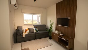 A modern living room with a grey sofa, a flat-screen TV mounted on a wooden panel, a potted plant, and a large window with a view of the outdoors. - APTO decorado 5 hospedes AC e piscina (Foz do Iguaçu)