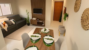 A modern living room with a sofa, TV, wooden cabinet, dining table with green placemats, and two chairs. - APTO decorado 5 hospedes AC e piscina (Foz do Iguaçu)