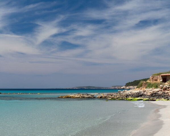 Ein Strand mit klarem türkisblauem Wasser, einer felsigen Küstenlinie und einem kleinen Gebäude auf einem Hügel.