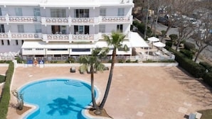 A hotel with a swimming pool, palm trees, and a multi-story building with balconies. - Apartment 5 In front of the sea (Urbanització Santo Tomàs, Es Migjorn Gran)