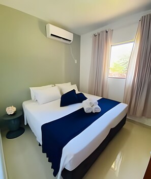 A neatly made bed with white and navy blue bedding, a small bedside table with a flower, and a window with curtains. - Apartamento para 4 pessoas com AC (Foz do Iguaçu)