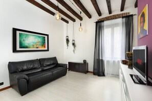 A modern living room with a black leather sofa, a framed abstract painting, hanging plants, a television, and a window with sheer curtains. - LECCO, 20 - In the heart of the Rainbow District. (Milan)
