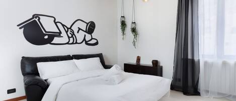 A modern bedroom with a black leather bed, white bedding, a wall-mounted light fixture, and a window with sheer curtains.