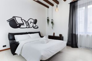 Um quarto moderno com uma cama de couro preta, roupa de cama branca, luminária fixada na parede e uma janela com cortinas leves.