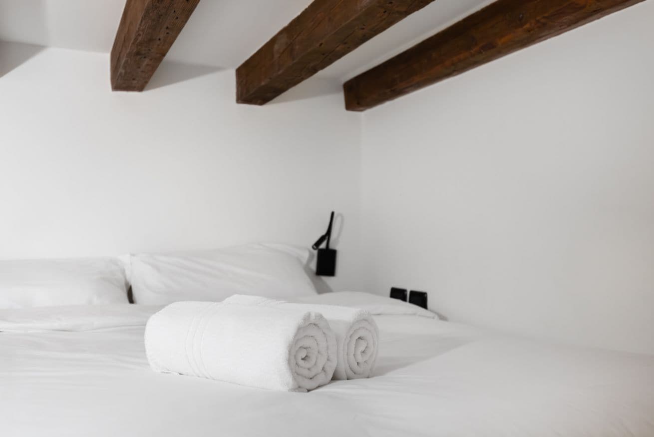 A white bed with rolled towels, wooden beams, a wall-mounted light, and a black object on the wall.