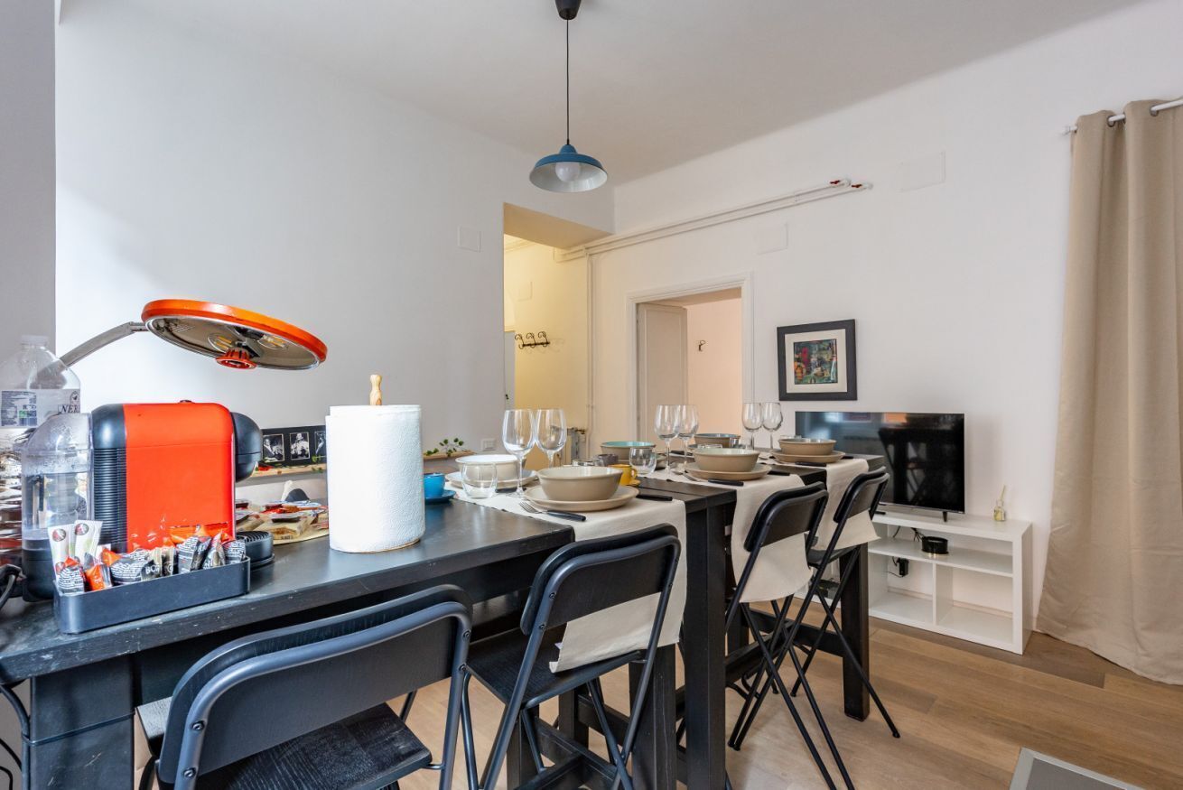 A modern kitchen with a dining table set for six, a coffee machine, and a TV in the background.