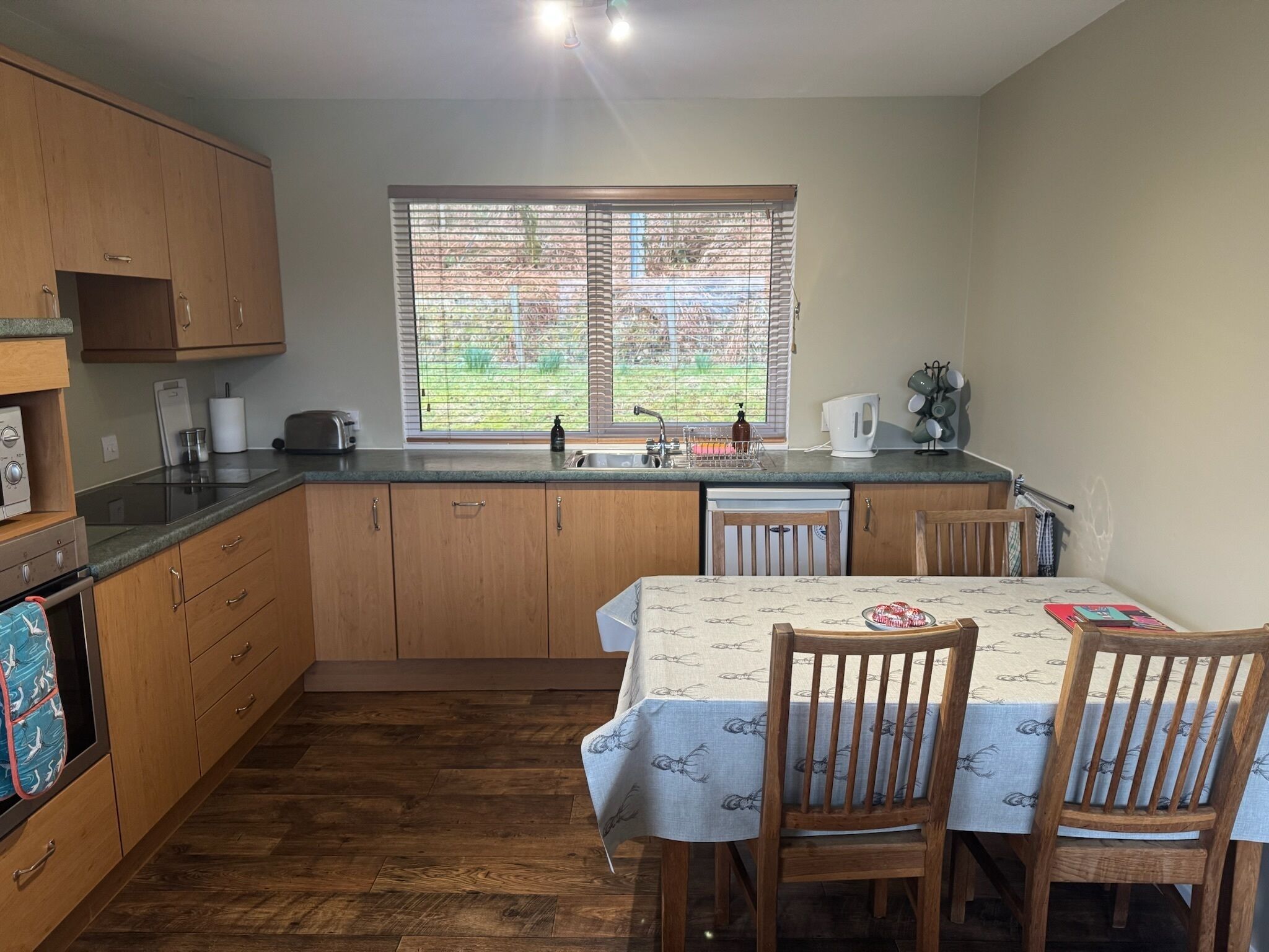 A kitchen with wooden cabinets, a dining table with chairs, a window with blinds, and a sink.