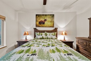 Cozy retreat with a large bed, bedside tables, and a dresser, featuring a forest-themed bedspread and a framed picture of a bear.