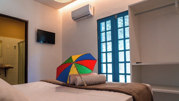 Une chambre d’hôtel avec un lit, un parapluie aux couleurs vives, une fenêtre avec des volets bleus et un téléviseur fixé au mur.