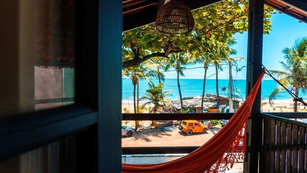 Blick auf den Strand von einer überdachten Veranda mit einer Hängematte, Palmen und einem parkenden Auto.